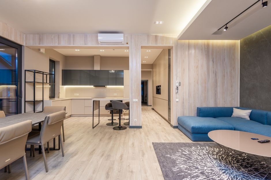 Interior of spacious modern studio apartment with comfortable blue corner sofa placed in front of dining table with chairs near kitchen zone